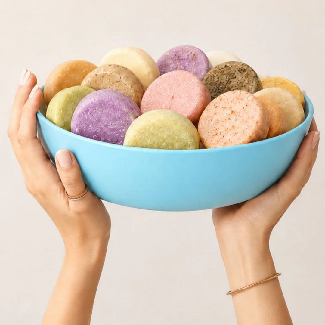 Hands holding a light blue bowl filled with assorted colorful conditioner bars.