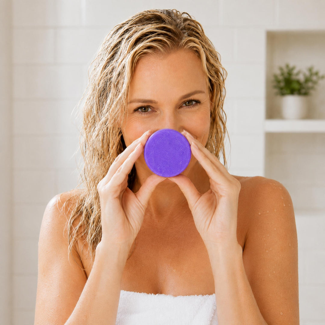 Woman with wet blonde hair holding a Dirty Bee Blondie Purple Conditioner Bar in a bright bathroom.