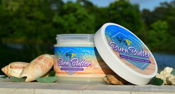 Dirty Bee Burn Butter body moisturizer displayed with seashells beside a lake.