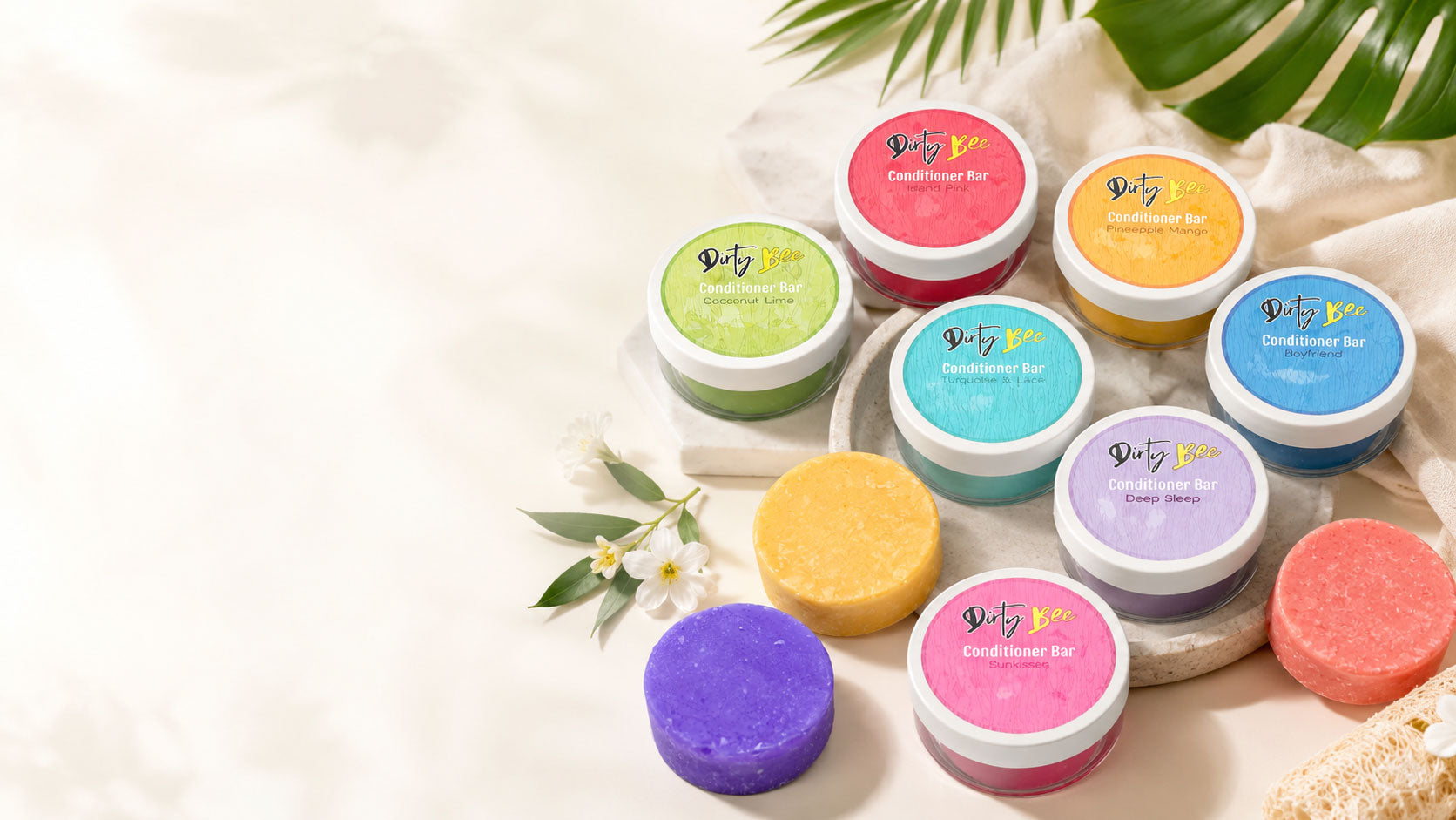 Assorted colorful Dirty Bee conditioner bars arranged with tropical leaves, flowers, and natural spa accessories.