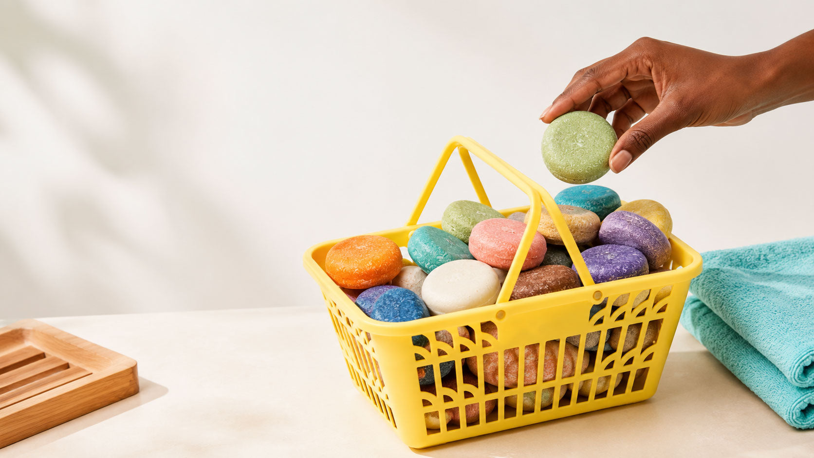 Yellow basket filled with colorful shampoo bars beside an aqua towel, with a hand lifting a green shampoo bar.